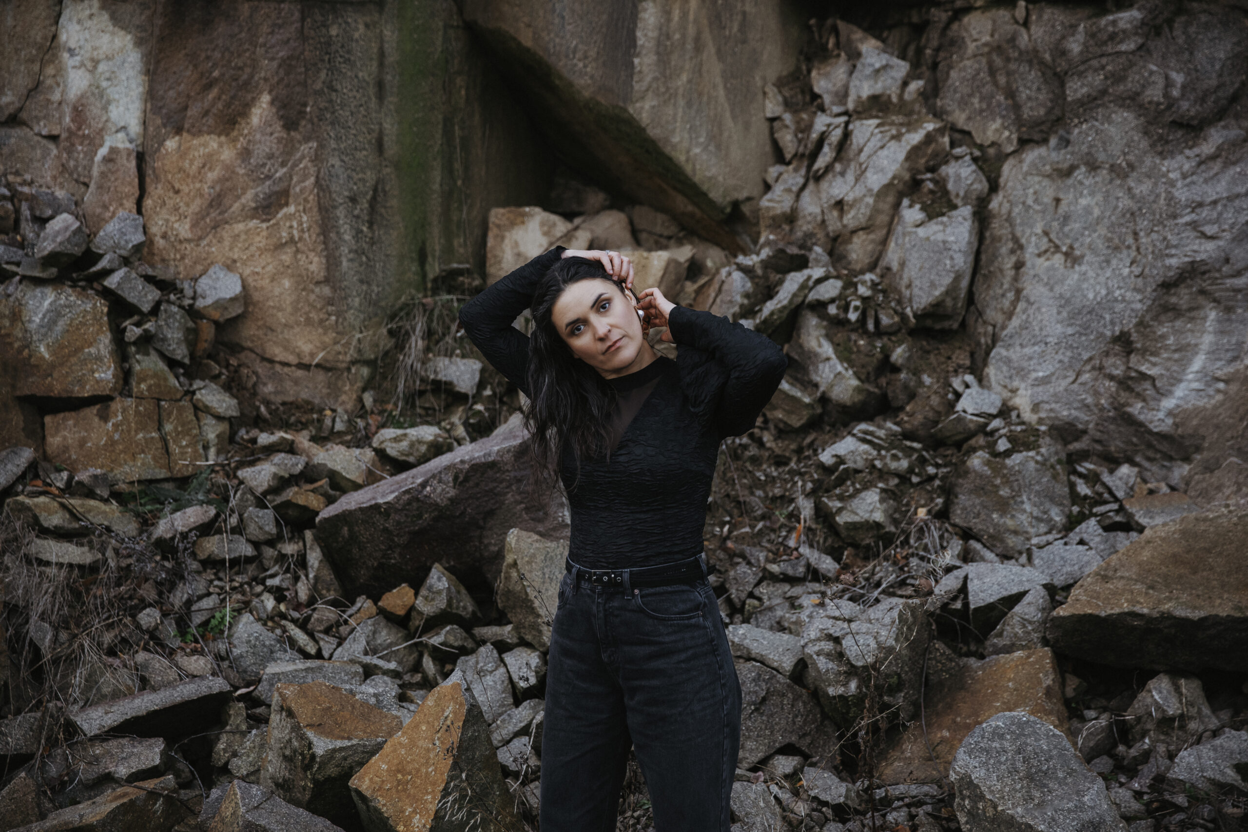 Eine Frau in schwarz gekleidet steht vor einem Steinbruch und fährt sich durch die braunen langen Haare