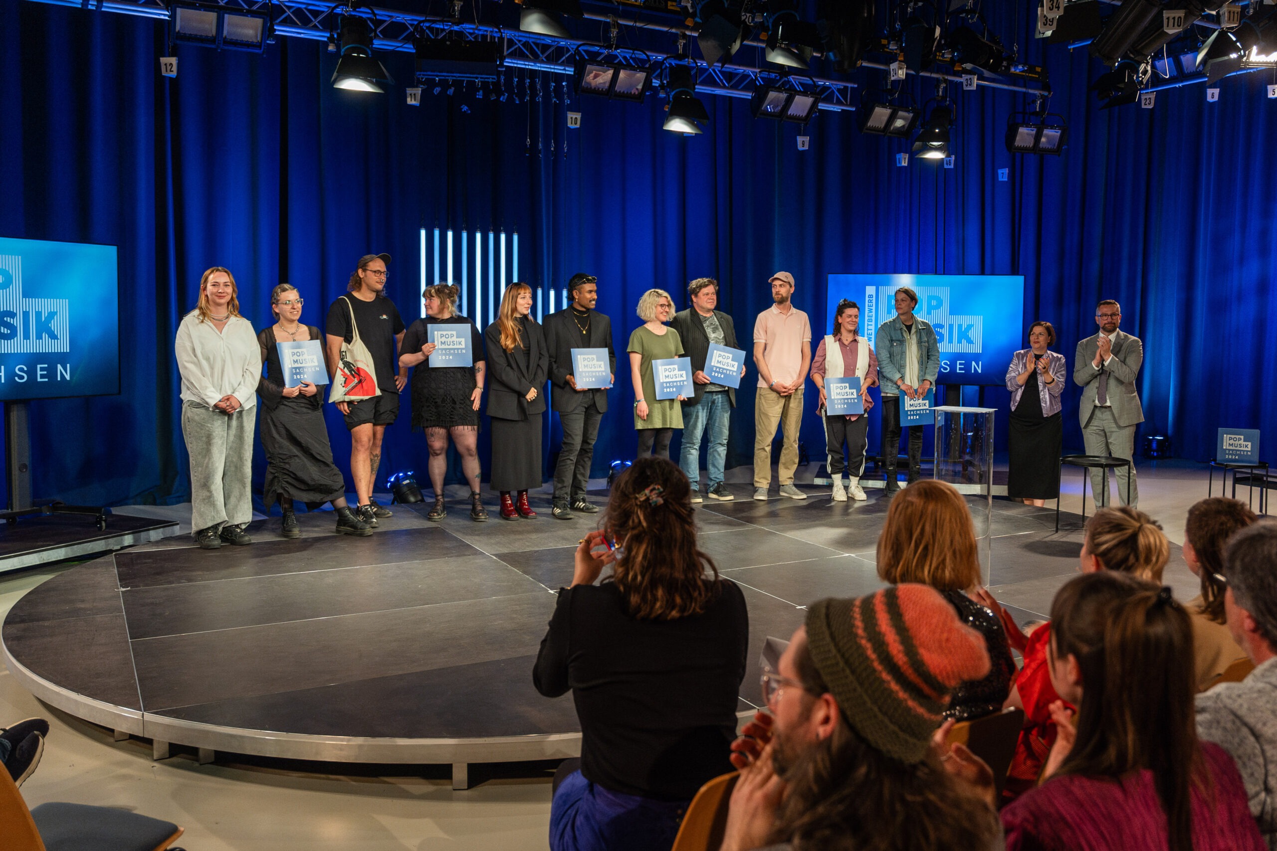 Eine Gruppe Menschen steht in Reihe auf einer Bühne in einem blau beleuchteten Fernsehstudio