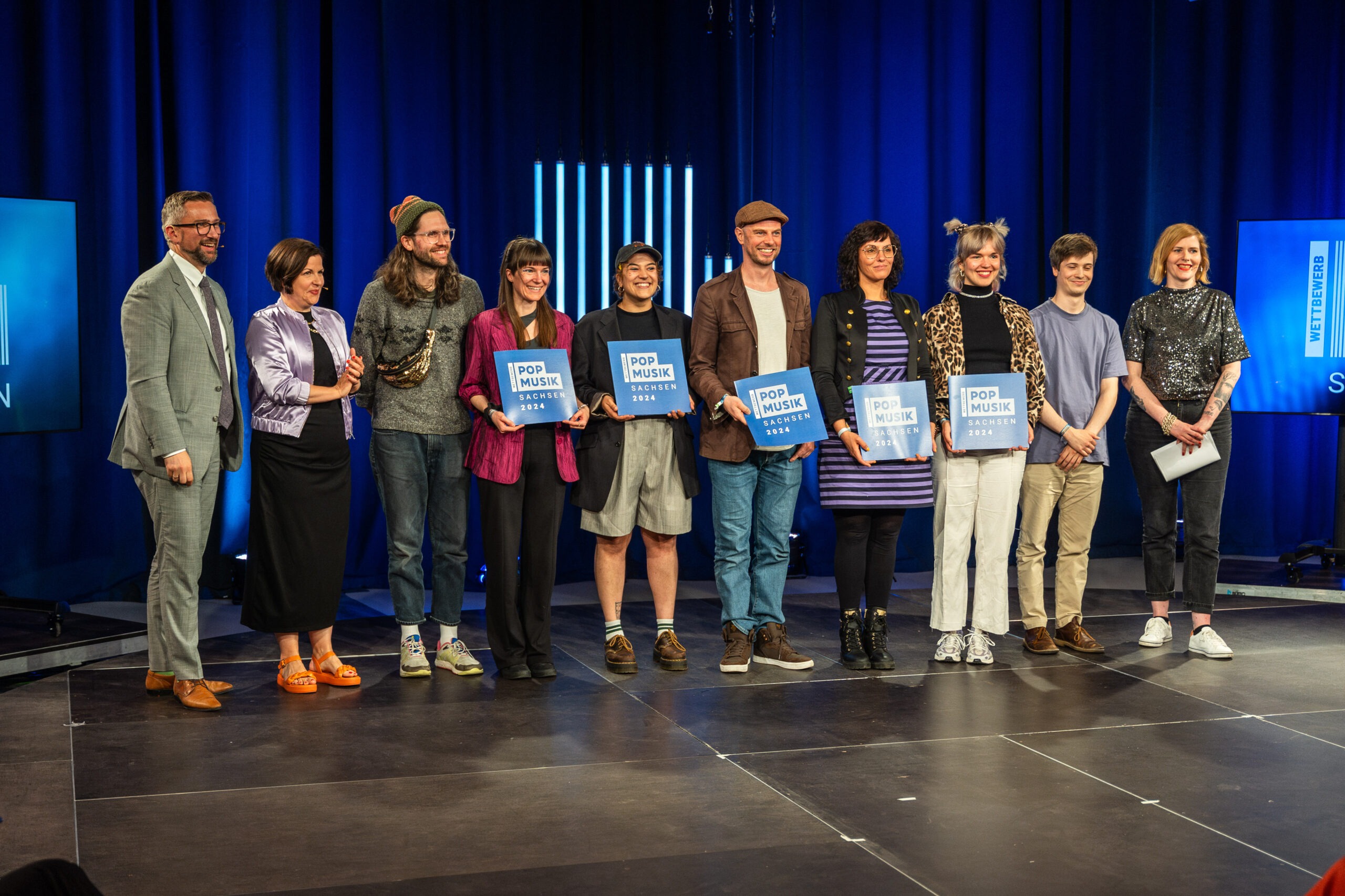 Eine Gruppe Menschen steht in Reihe auf einer Bühne in einem blau beleuchteten Fernsehstudio