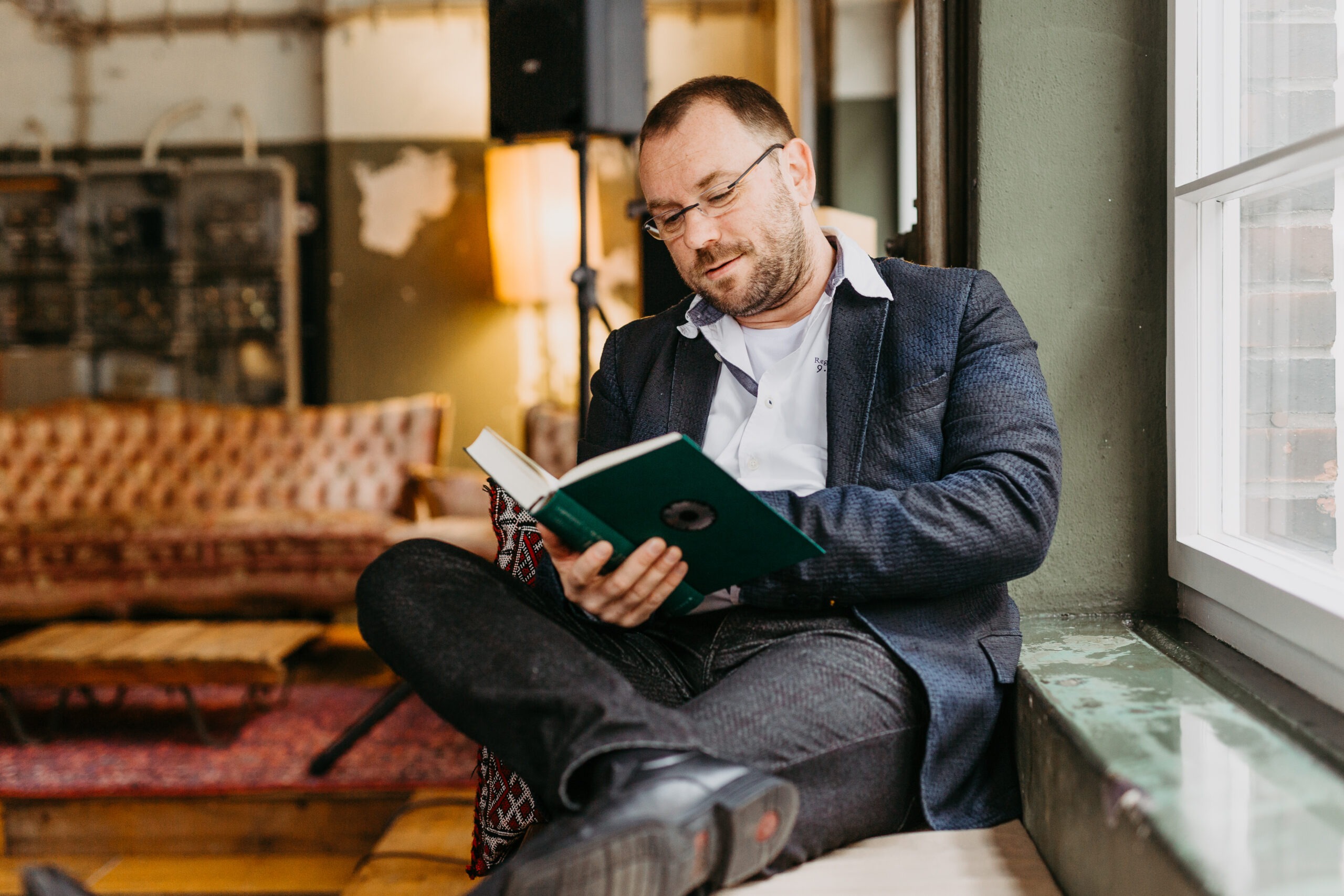 Mann mit kurzen Haaren und Brille liest in einem Buch. Er hat das rechte Bein übergeschlagen und sitzt an einem Fenster.