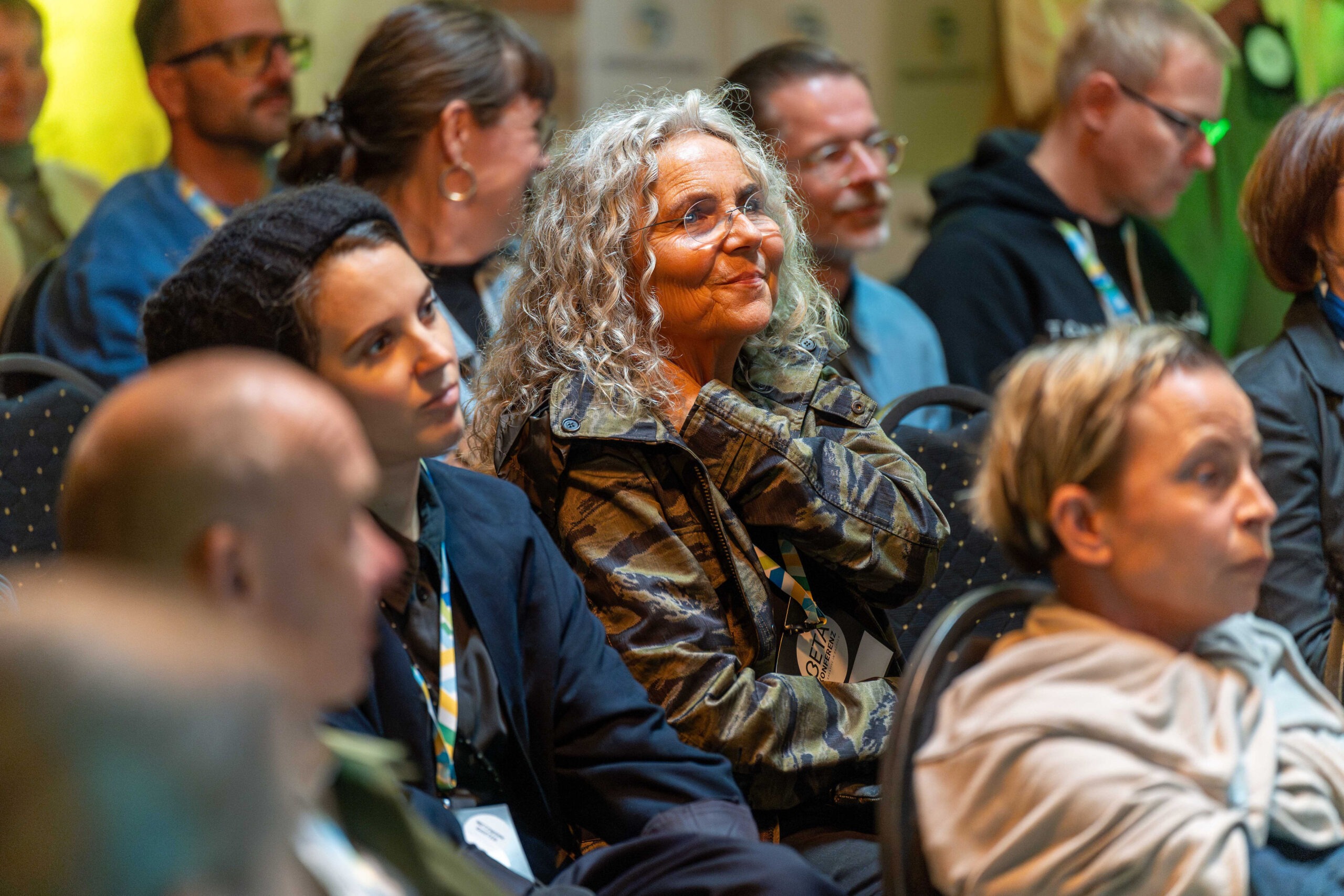 Eine Frau mit grauen Locken und Brille sitzt im Publikum zwischen anderen Menschen und schaut nach vorne. Sie lächelt.