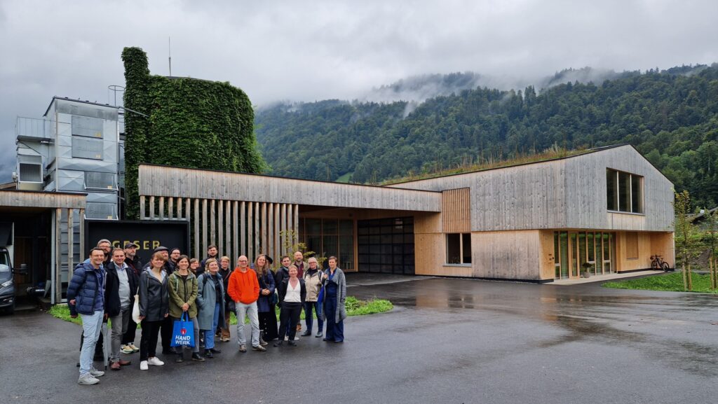 Gruppenbild von 20 Menschen im Regen vor einem großen Holzhaus, im Hintergrund Nebel und Wälder