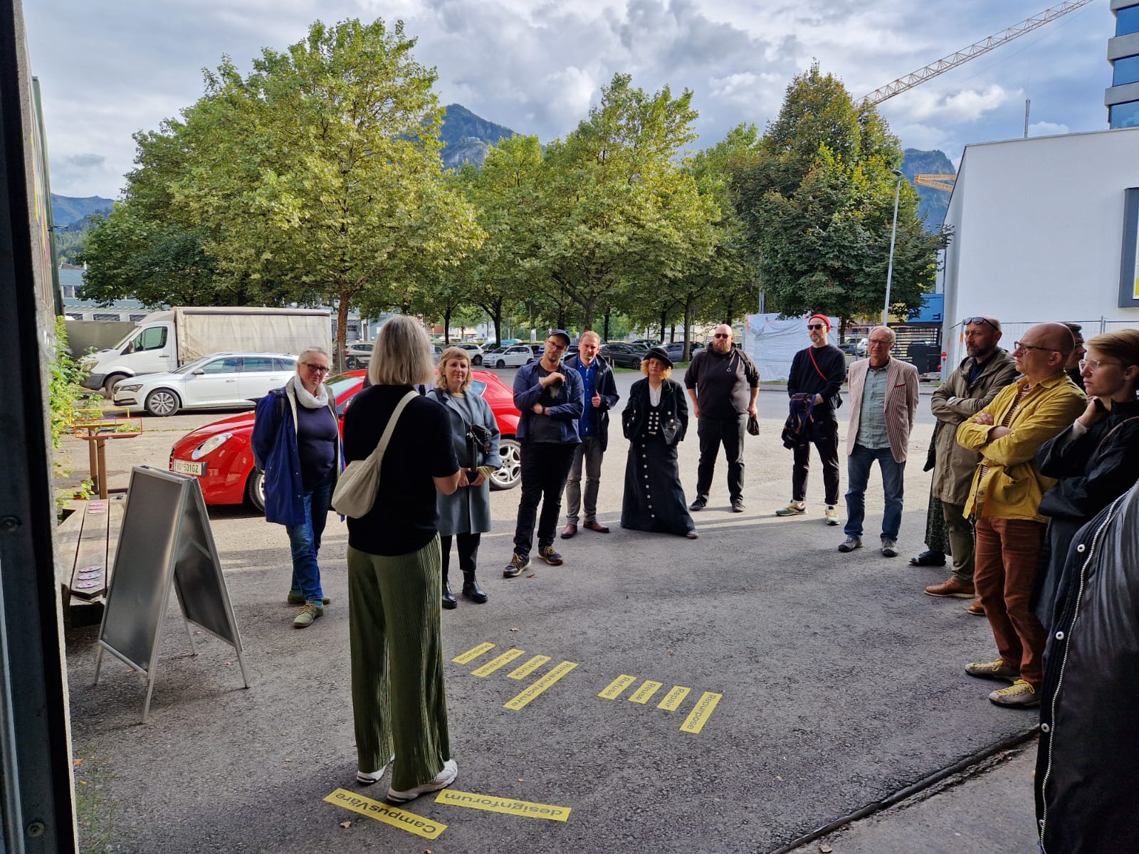 Menschen stehen auf einer Straße im Halbkreis und unterhalten sich, im Hintergrund grüne Bäume.