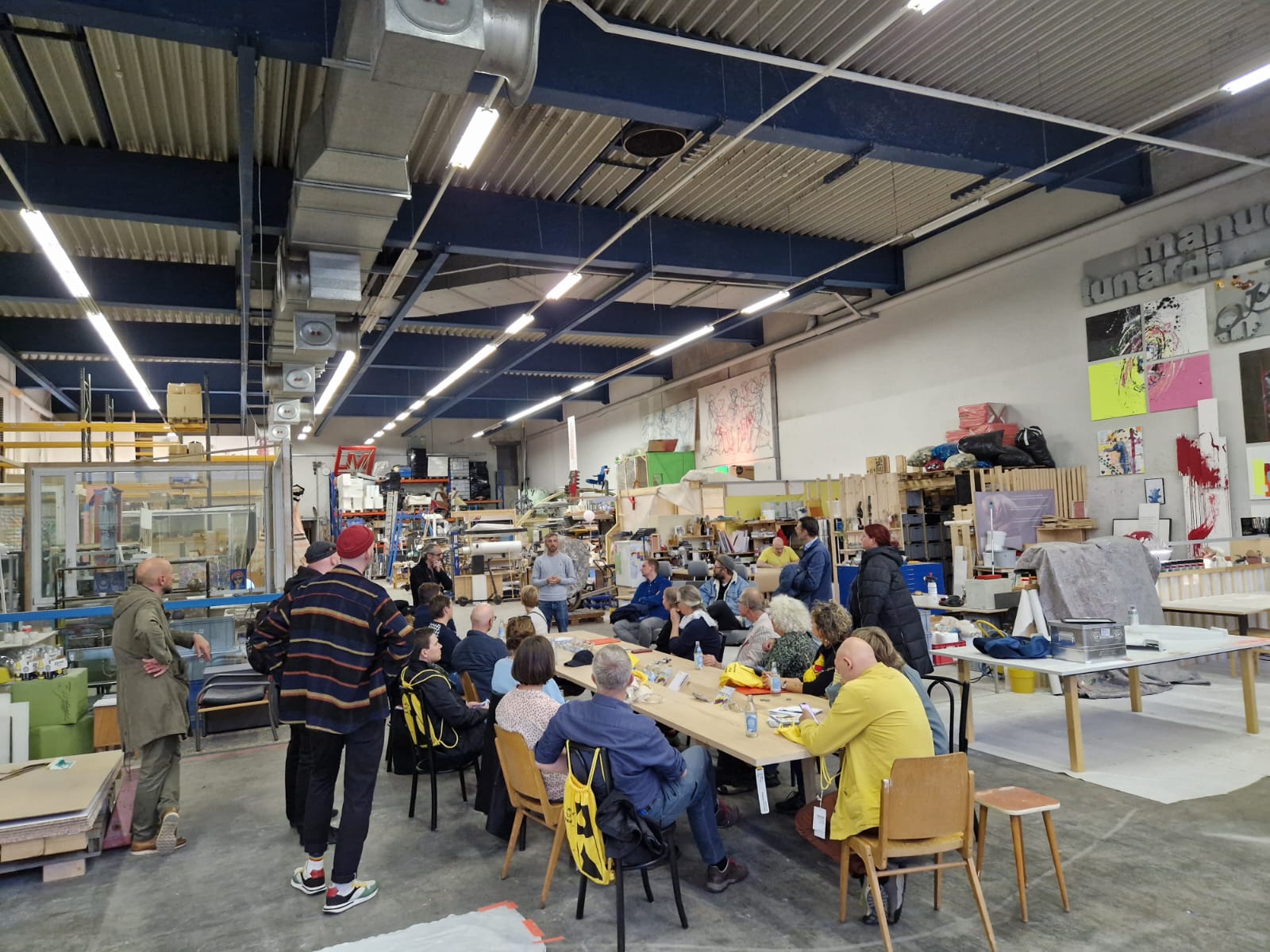 Eine Gruppe von 20 Menschen sitzt an einem großen Tisch mitten in einem Atelier. An der Decke industrielle Lampen.