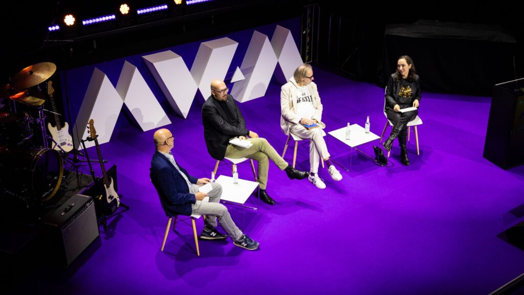 Vier Menschen sitzen auf einer Bühne mit lila Boden, das Foto ist von oben aufgenommen. Im Hintergrund die Schrift: MWM